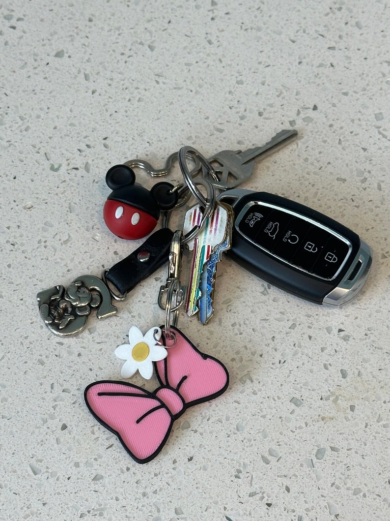 May include: A set of keys with a pink bow keychain, a white daisy charm, a silver Mickey Mouse charm, and a black car key with a silver key ring.