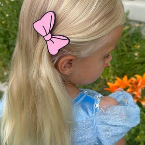 May include: A young child with long, blonde hair wearing a light blue dress and a pink bow hair clip. The hair clip is a solid pink color with a black outline. The child is outdoors with flowers in the background.