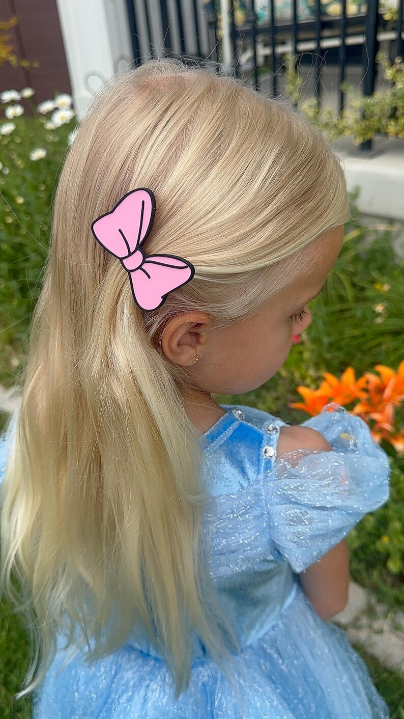 May include: A young child with long, blonde hair wearing a light blue dress and a pink bow hair clip. The hair clip is a solid pink color with a black outline. The child is outdoors with flowers in the background.