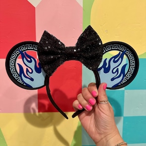 May include: Black sequined bow headband with round ear accents featuring a blue flame design. The ears have a black border with a Greek key pattern. The headband is black and held up by a hand with pink nail polish.