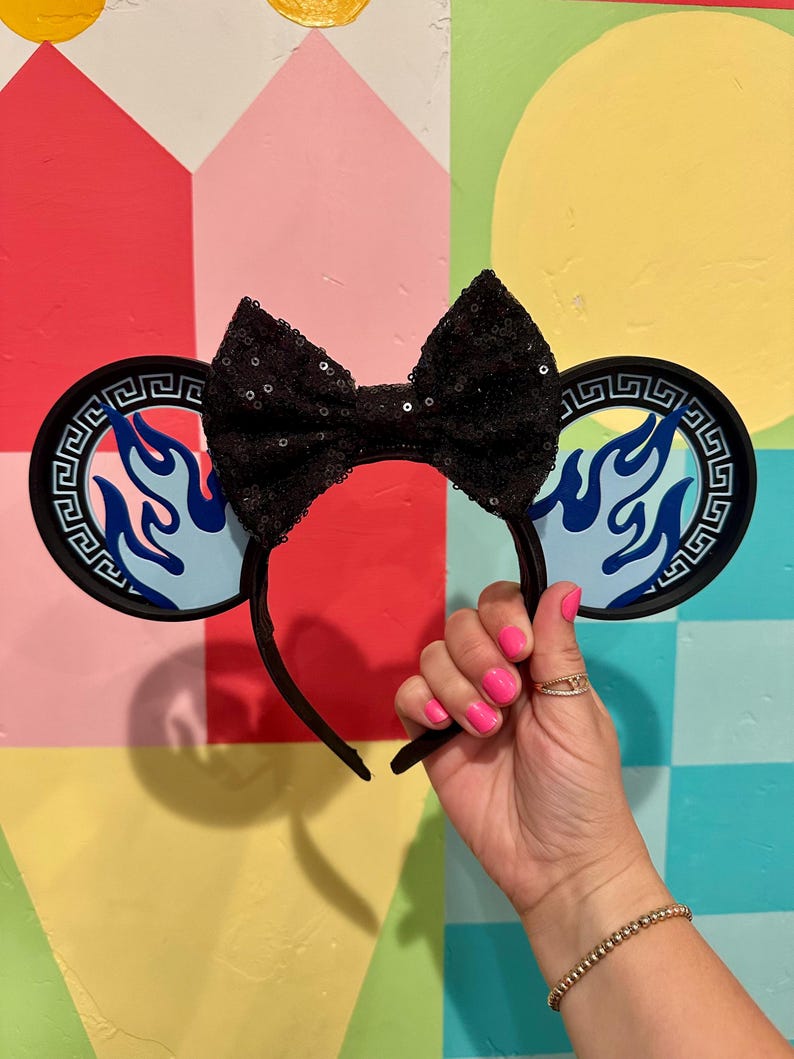 May include: Black sequined bow headband with round ear accents featuring a blue flame design. The ears have a black border with a Greek key pattern. The headband is black and held up by a hand with pink nail polish.