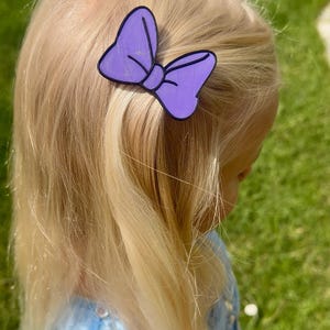 May include: A purple bow-shaped hair clip with a black outline is attached to blonde hair. The hair clip is a solid purple color with vertical lines. The background is blurred, showing green grass and a blue dress.
