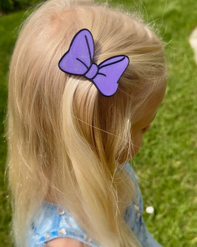 May include: A purple bow-shaped hair clip with a black outline is attached to blonde hair. The hair clip is a solid purple color with vertical lines. The background is blurred, showing green grass and a blue dress.