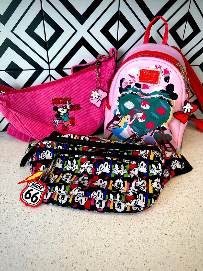 May include: Three pink and black Disney-themed bags. The largest bag is a pink backpack with a red zipper and a white Alice in Wonderland design. The middle bag is a pink corduroy shoulder bag with a Mickey Mouse design. The smallest bag is a black fanny pack with a colorful Mickey Mouse design.