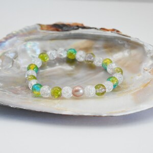 May include: A white, green, and blue crystal bead bracelet with a single pink pearl bead. The bracelet is displayed on a white seashell.