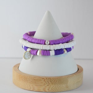 May include: Three layered bracelets with purple, white, and light purple beads. The bracelets have a gold charm with a smiley face on it. The bracelets are displayed on a white cone-shaped stand with a wooden base.