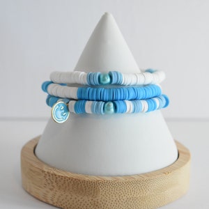 May include: Stack of three beaded bracelets in shades of blue and white, displayed on a white cone-shaped stand with a wooden base. One bracelet features a gold charm with a blue smiley face.