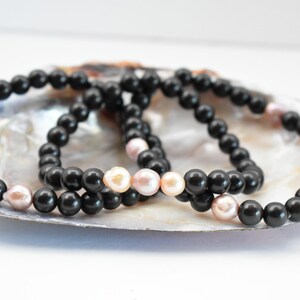 Puede incluir: Tres pulseras de cuentas negras con detalles de perlas rosadas. Las pulseras se muestran sobre una concha de nácar blanca.