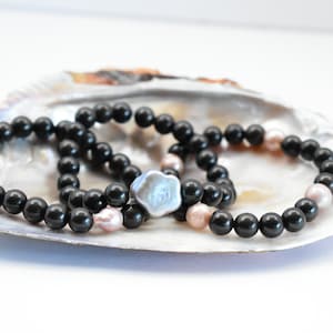 May include: Two black beaded bracelets with a single large pearl charm, displayed on a white seashell.