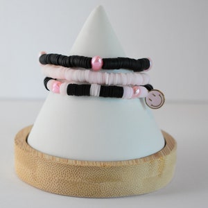 May include: Three layered bracelets with black and white beads and pink accents. The bracelets are stacked on a white cone-shaped stand with a bamboo base. A pink smiley face charm is attached to the bottom bracelet.