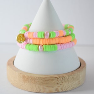 May include: A stack of three bracelets made with colorful beads. The bracelets are pink, orange, and green. The bracelets are on a white cone-shaped stand with a wooden base.