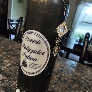 Puede incluir: Una botella negra con una etiqueta que dice "Premade Polyjuice Potion". La botella está adornada con alambre y adornos de joyas. La etiqueta incluye instrucciones para agregar un pedazo de la persona cuya apariencia física desea asumir.