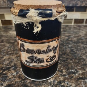 Puede incluir: Un tarro cilíndrico negro con un tapón de corcho y una etiqueta que dice "Boomslang Skin". El tarro está envuelto con hilo y tela blanca. La etiqueta tiene un borde marrón y el texto es de estilo vintage.
