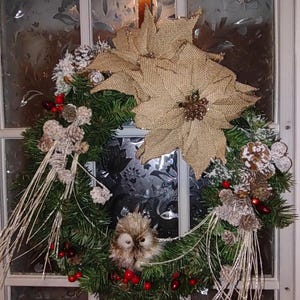 Puede incluir: Una corona de Navidad con una poinsettia de arpillera, ramas de pino, bayas rojas, piñas blancas y una pequeña figura de búho marrón.