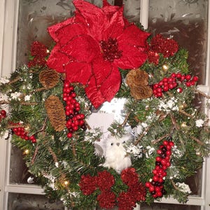 Puede incluir: Una corona de Navidad con una flor de pascua roja, bayas rojas, ramas de pino y un adorno de búho blanco.