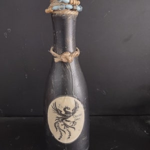 Op de afbeelding: Een zwarte glazen fles met een kurk en een bruine touw strik. De fles heeft een wit etiket met een zwart phoenix ontwerp en de tekst "Phoenix Tears".