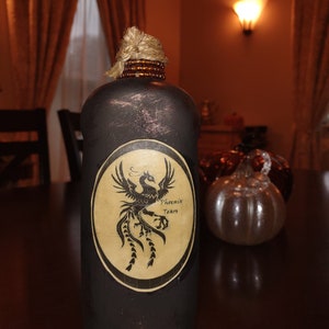 Op de afbeelding: Een zwarte glazen fles met een bruine kurkstop en een etiket met de tekst "Phoenix Tears" en een zwart silhouet van een feniks. De fles staat op een donker houten oppervlak.