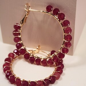 May include: Gold hoop earrings adorned with faceted, deep red beads. The beads are strung along the gold hoops, creating a continuous circle. The earrings are displayed against a soft, neutral background.