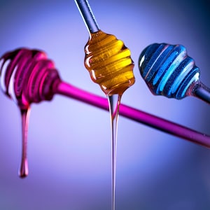 May include: Three honey dippers, one with honey dripping from it. The dippers are pink, blue, and clear with a yellow honey drizzle.