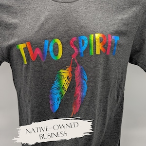 Könnte beinhalten: Dunkelgraues T-Shirt mit den Worten „TWO SPIRIT“ in Regenbogenfarben. Darunter befinden sich zwei regenbogenfarbene Federn. Das Shirt trägt auch den Text „NATIVE-OWNED BUSINESS“ in einem weißen Banner.