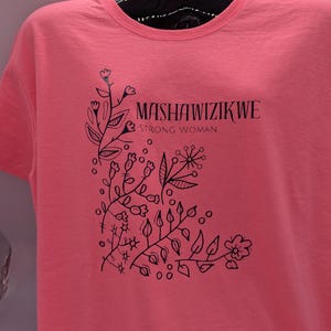 Peut inclure: T-shirt corail avec un motif floral noir dans le coin et les mots "MASHAWIZIKWE STRONG WOMAN". Le motif comprend des feuilles, des fleurs et des points. Le t-shirt a un col rond et des manches courtes.