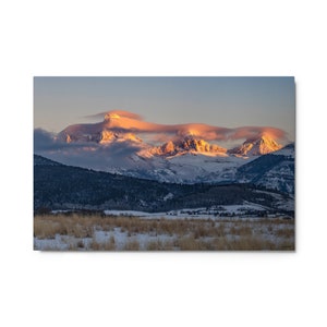 May include: A panoramic view of snow-capped mountains with a layer of clouds illuminated by the golden light of sunrise. The mountains are covered in snow and the foreground is a field of dry grass.