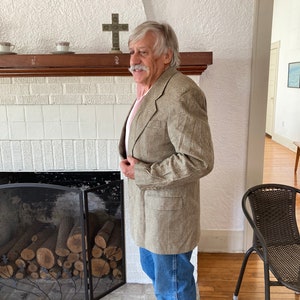 Puede incluir: Un hombre con un blazer de cuadros marrones y blancos con una camisa rosa claro debajo. Está de pie frente a una chimenea con una pantalla de metal negro y troncos en su interior. Hay una repisa de madera sobre la chimenea con una cruz encima.