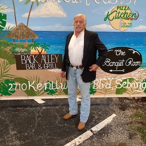 Puede incluir: Un hombre con un blazer negro y pantalones vaqueros azules se encuentra frente a un mural que representa una escena de playa tropical. El mural incluye el texto "Downtown Oasis!" y "Back Alley Bar & Grill".
