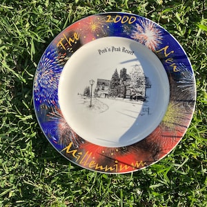 May include: A white ceramic plate with a black and white illustration of a building with a gold inscription that reads "The New Millennium 2000 Peek'n Peak Resort". The plate has a blue and red border with gold fireworks.
