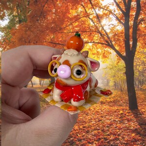 May include: A small, cream-coloured cow figurine with brown spots, wearing round yellow glasses and a bright red scarf. A miniature orange pumpkin sits on its head. The cow rests on a small, square, yellow and white checkered blanket.