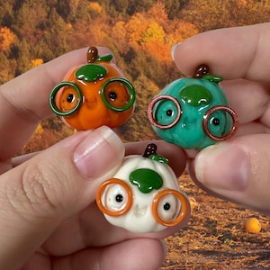 May include: Three small, smooth, rounded pumpkin figurines made of glossy material. One is orange, one is teal, and one is white. Each pumpkin features a green stem, a small smiling face, and a pair of round glasses in a contrasting colour.