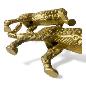 May include: Two gold-colored cheetah figurines are positioned on a white background. The cheetahs are in a running pose, with detailed textures and visible spots. The figurines are made of a metallic material.