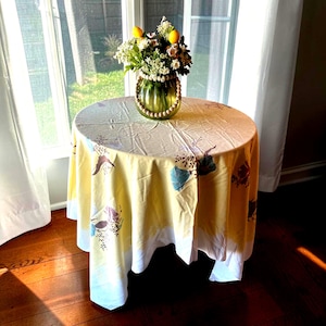 May include: A round table covered with a yellow and white tablecloth featuring a floral pattern. A green glass vase with a wooden bead handle sits on the table, filled with a bouquet of white flowers and two yellow lemon-shaped decorations.