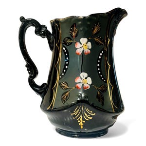 May include: A dark green ceramic pitcher with a black handle and spout. The pitcher is decorated with white flowers with red centers, gold accents, and gold leaf designs. The pitcher has a capacity of approximately 64 ounces.