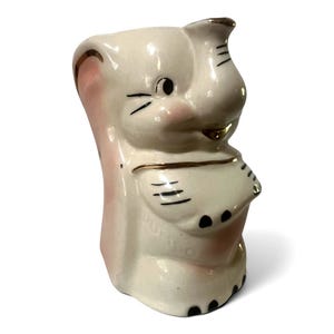 May include: A cream-colored ceramic creamer shaped like an elephant with pink accents on the ears and cheeks. The piece features gold-painted trim along the ears, trunk, and body, with black painted details for the eyes, toes, and facial features.