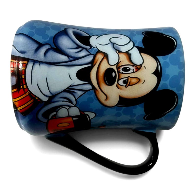 May include: A ceramic mug featuring a distressed Mickey Mouse design. The mug has a black handle and a blue background with a pattern of Mickey Mouse silhouettes. Mickey is depicted with a tired expression, wearing a light blue jacket.
