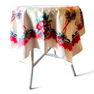 May include: A round tablecloth with an off-white base featuring a repeating pattern of red and blue floral bouquets connected by red ribbons and blue twisted rope motifs, finished with a scalloped red border along the bottom edge of the fabric.