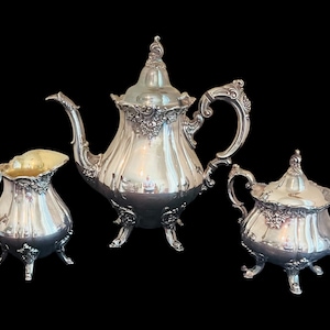 May include: A silver tea set with ornate floral detailing. The set includes a teapot, creamer, and sugar bowl, all with curved handles and decorative feet. The teapot has a tall, curved spout and a domed lid. The creamer has a gold interior.