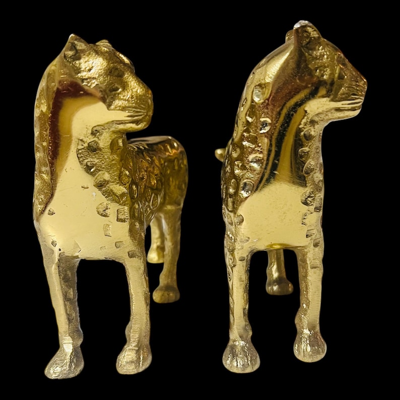 May include: Two gold-colored metal horse figurines. The horses are standing, with textured surfaces and detailed features. The background is black, highlighting the figurines' metallic sheen. The horses are facing different directions.