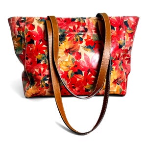 May include: A tote bag featuring a vibrant floral pattern with shades of red, orange, and yellow on a cream background. The bag includes two long, smooth brown leather shoulder straps with visible white stitching along the edges of the straps.