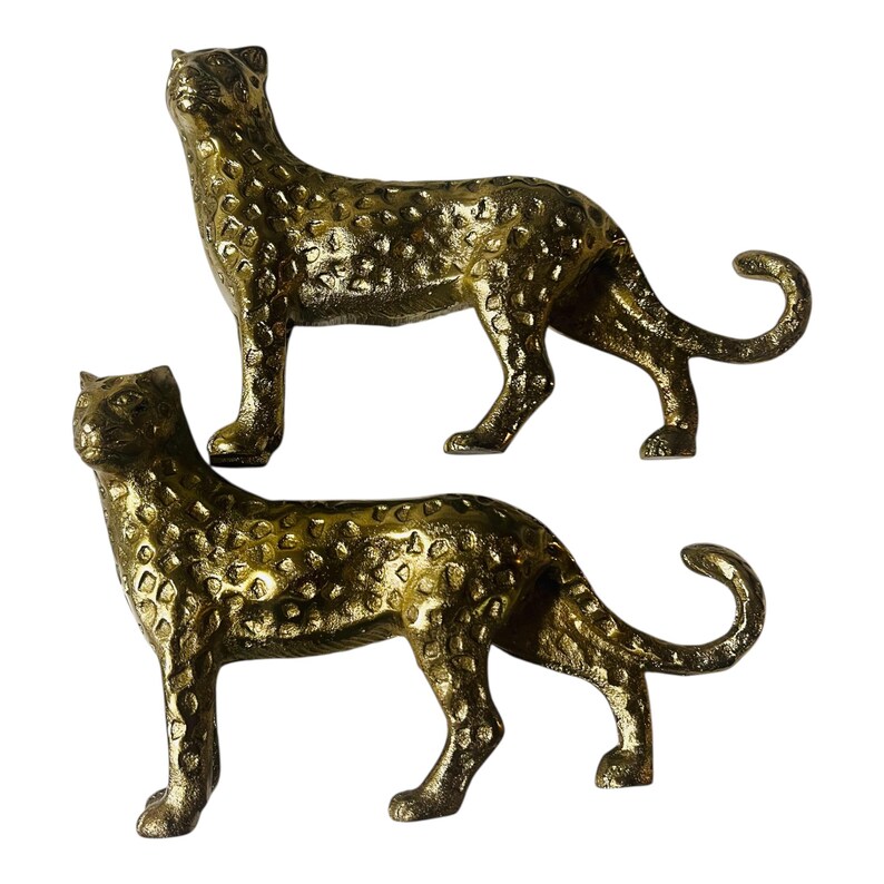 May include: Two gold-colored leopard figurines. The figurines are in a standing pose with their tails curled. The surface of the leopards has a textured, spotted pattern. The figurines are made of a metallic material.