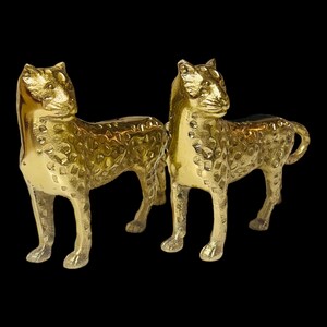 May include: Two gold-colored, textured sculptures of big cats, possibly cheetahs or leopards, are displayed against a black background. The statues are detailed with spots and stand in a poised position, suggesting movement.