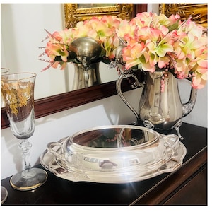May include: A silver serving dish with a lid and handles, resting on a matching tray. A tall, ornate champagne glass with gold detailing is on the left. A silver pitcher with pink and yellow flowers is in the background.