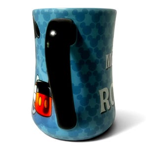 May include: A light blue ceramic mug featuring a pattern of Mickey Mouse silhouettes. The mug has a black handle and a partial image of Mickey Mouse. The word "Mickey" is visible in white text on the side of the mug.