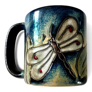 May include: A ceramic mug with a dragonfly design. The mug has a dark blue and green glaze with a black handle. The dragonfly is light gray with red accents. The mug is approximately 4 inches tall.