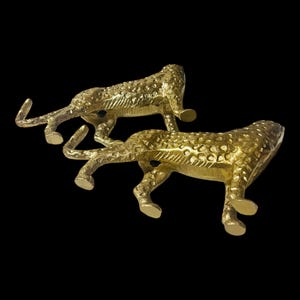 May include: Two gold-colored metal cheetah figurines. The cheetahs are in a walking pose, with detailed spots and textured surfaces. The background is solid black.