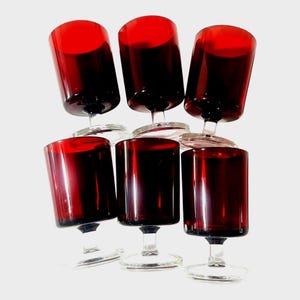 May include: A set of six matching drinking glasses with deep red bowls and clear glass stems and circular bases. Each glass features a cylindrical bowl shape with a smooth finish, arranged in two rows of three against a plain white background.