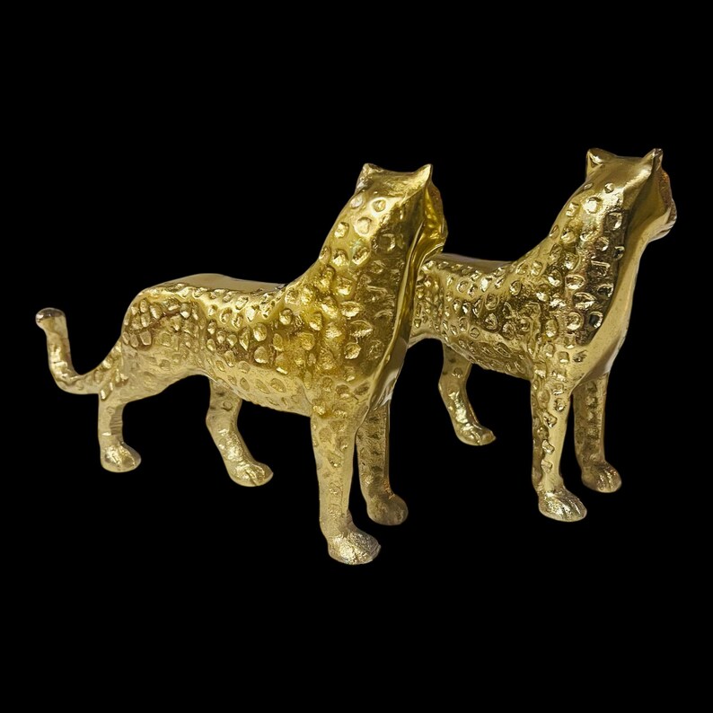 May include: Two gold-colored cheetah figurines with textured surfaces and spotted patterns. The cheetahs are standing side-by-side, facing the same direction, with their tails curled. The background is black.