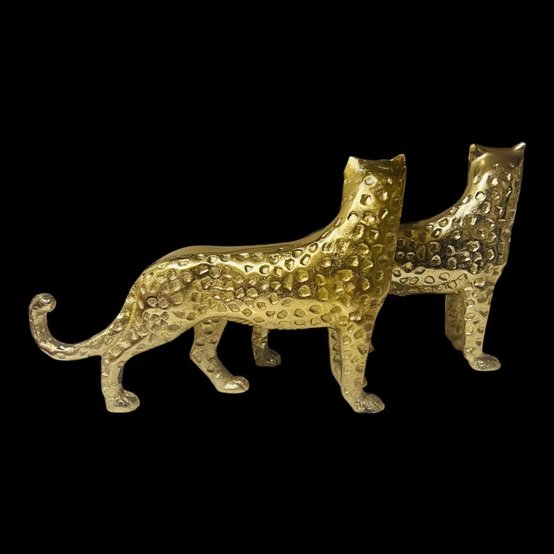May include: Two gold-colored leopard figurines with textured surfaces. The leopards are in a standing pose, with their tails curled. The background is solid black, highlighting the metallic finish of the sculptures. These decorative pieces could be used as home decor.