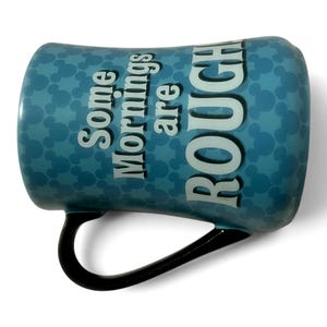 May include: A blue ceramic mug with a black handle. The mug features a pattern of small circles and the text "Some Mornings are ROUGH" in white. The mug is designed for hot beverages.